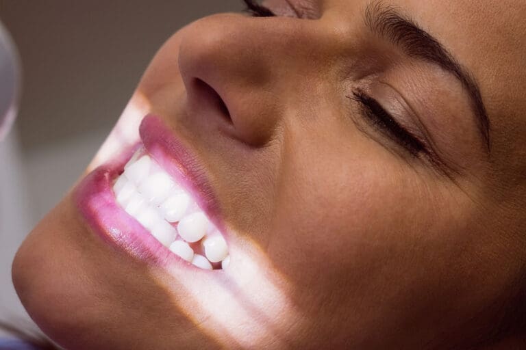 A close-up of a smiling person with bright white teeth and smooth skin, illuminated by a soft beam of light across their face. Their eyes are closed, and they appear relaxed and happy, showcasing the results from a Lansing dentist.