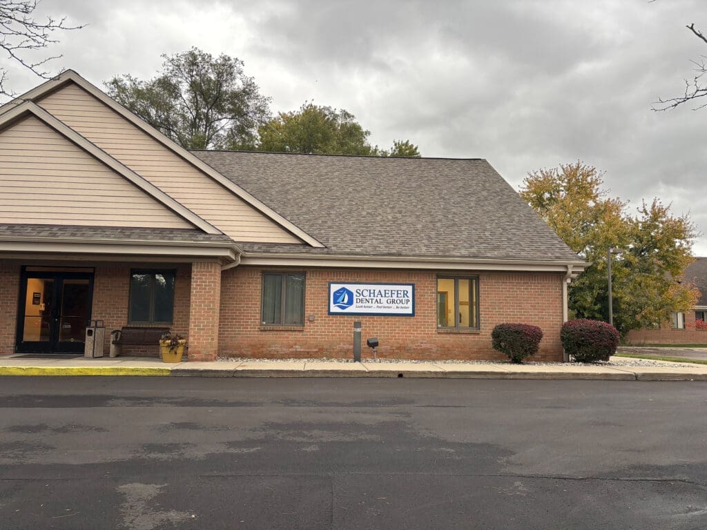A one-story brick dental clinic with a sign reading Schaefer Dental Group stands near shrubs by the entrance under cloudy skies. The parking lot is empty.