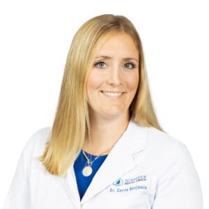 A woman with straight blonde hair, wearing a white lab coat that reads Schaefer Dental Group, Dr. Carrie Benjamin, and a blue top, smiles at the camera. She represents a trusted Lansing dentist in a dental clinic setting. The background is plain white.