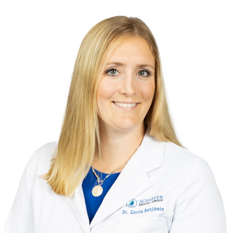 A woman with straight blonde hair, wearing a white lab coat that reads Schaefer Dental Group, Dr. Carrie Benjamin, and a blue top, smiles at the camera. She represents a trusted Lansing dentist in a dental clinic setting. The background is plain white.