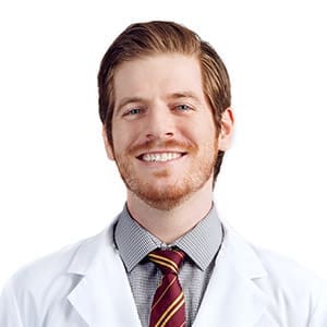 Smiling man with light brown hair and beard, wearing a white lab coat, checkered shirt, and striped tie, posing in front of a plain white background.