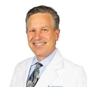 A smiling middle-aged man with short, grayish hair wears a white lab coat, blue shirt, and patterned tie, standing against a plain white background—an approachable Lansing dentist ready to welcome you to his dental clinic.