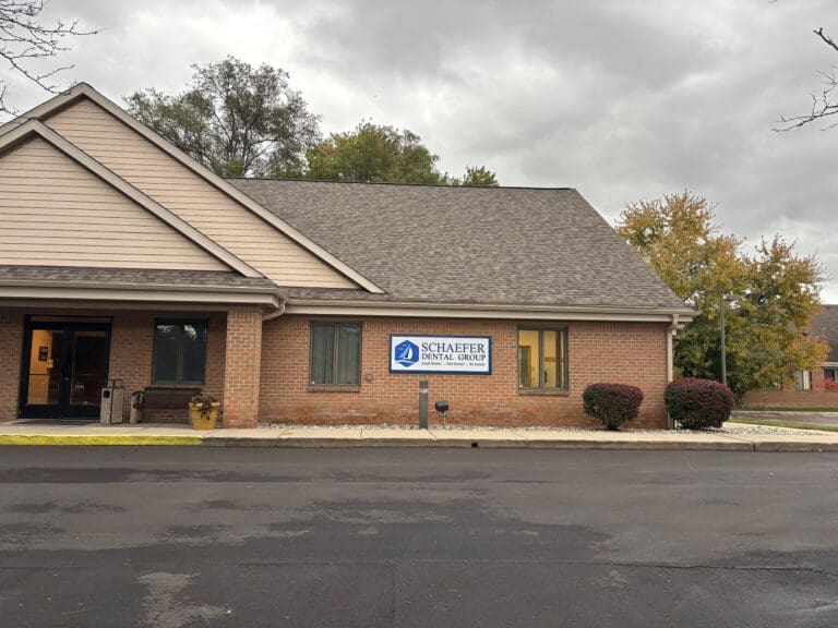 A one-story brick dental clinic with a sign reading Schaefer Dental Group stands near shrubs by the entrance under cloudy skies. The parking lot is empty.